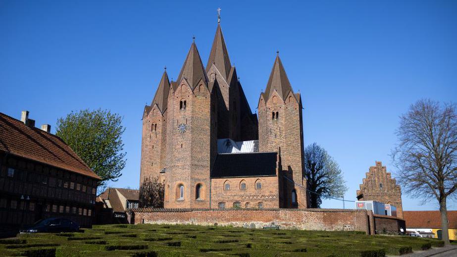 Brick church towers in striking Danish architectural style stand against a backdrop of blue sky with trees and neatly cut hedges in the foreground.