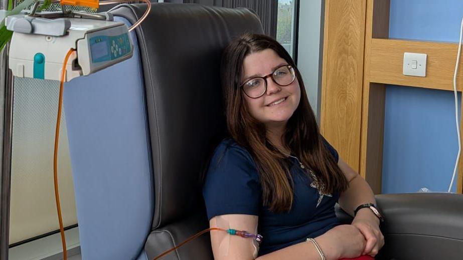 Daisy yn eistedd mewn cadair ysbyty yn derbyn triniaeth cemotherapi, gyda chyfarpar meddygol o’i chwmpas