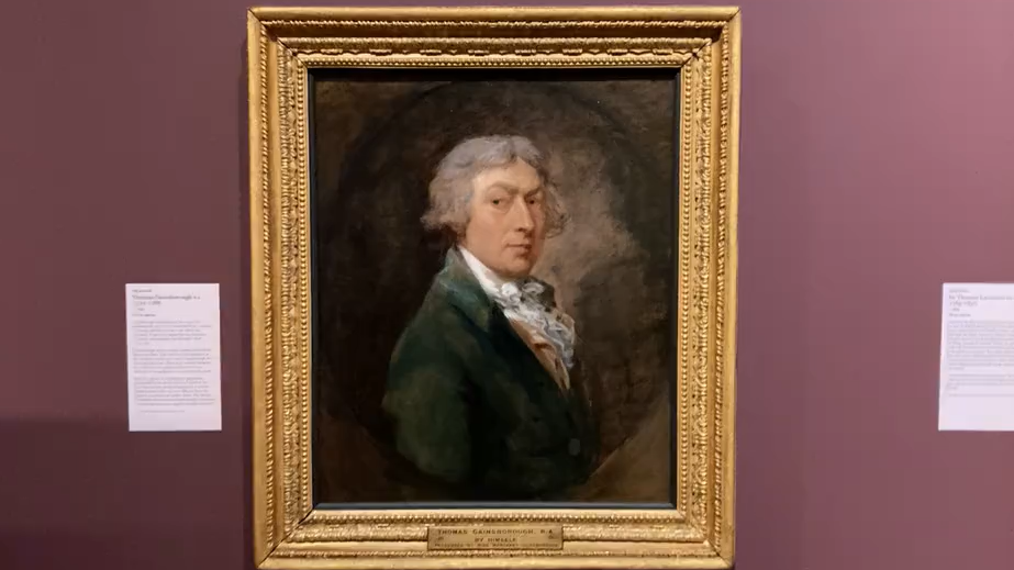 An oil portrait in an ornate gold frame on a dark pink wall. It is of a grey-haired Thomas Gainsborough in right-side profile, looking to the right. He is dressed in a dark green jacket, with a white ruffle-coloured shirt 