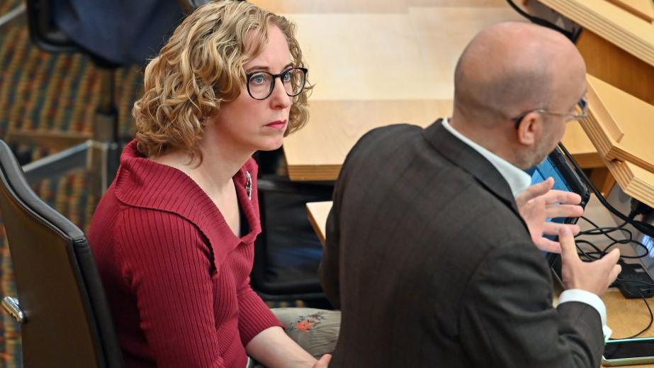 Lorna Slater, with blonde curly hair and black glasses sits in a chair in Holyrood, she is looking up a Patrick Harvie. He is bald with glasses but on the back of his head is visible. He is mid speech and is gesturing with his hands.