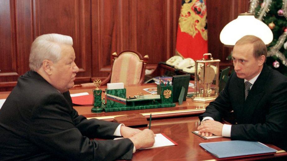 Boris Yeltsin sits opposite Vladimir Putin at a table, looking at each other, the day Yeltsin annouced he was stepping down as president