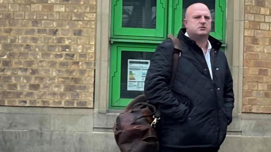 Michael Kempson: A bald man standing in front of brown-brick wall. He has his hands in the pockets of a black, zipped, jacket and is carrying a brown bag over his right shoulder.