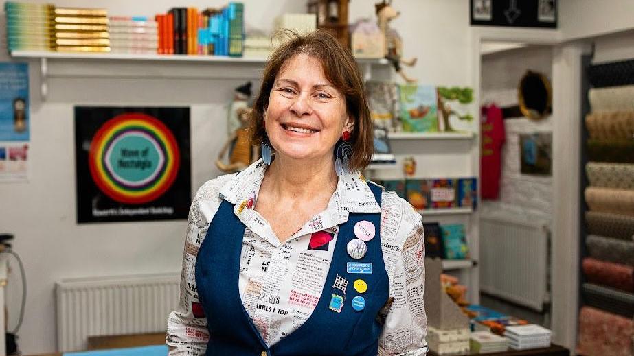 A woman with short brown hair wearing a white shirt with printed text on it and a navy blue waistcoat with badges on. She is standing in a bookshop. 