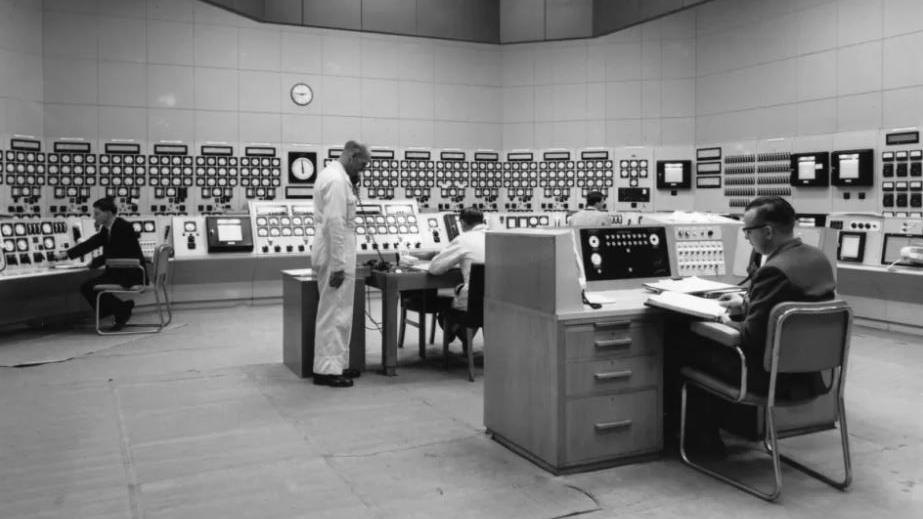 A black and white image showing a control room inside the Berkeley nuclear power station. There are men sat behind desks making notes, and another man in white overalls standing up on the phone. The walls are covered in circular dials, buttons and valves. 