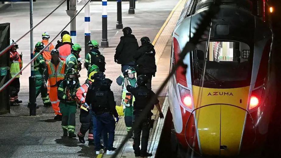A yellow and red and white train parked on the station, with emergency workers standing next to it on the platform.