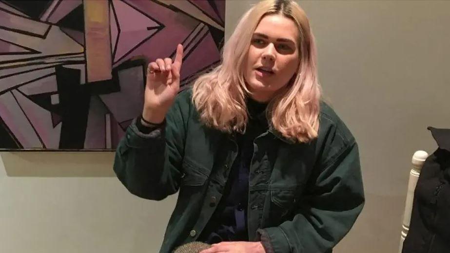 Bethan Roper sits in front of a large pop art picture. She is wearing a green shirt jacket, and dark top. she is holding her hand up. She has pink-tinged blond hair and dark eyebrows. 