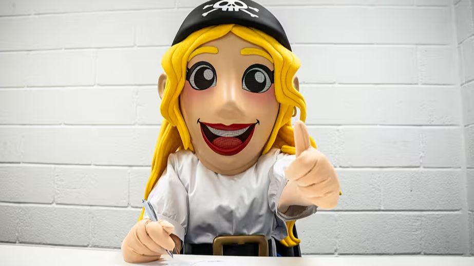 The new Bristol Rovers mascot Irene sits at a table and gives a thumbs up to the camera. She is in a puppet outfit with yellow hair and a big smile