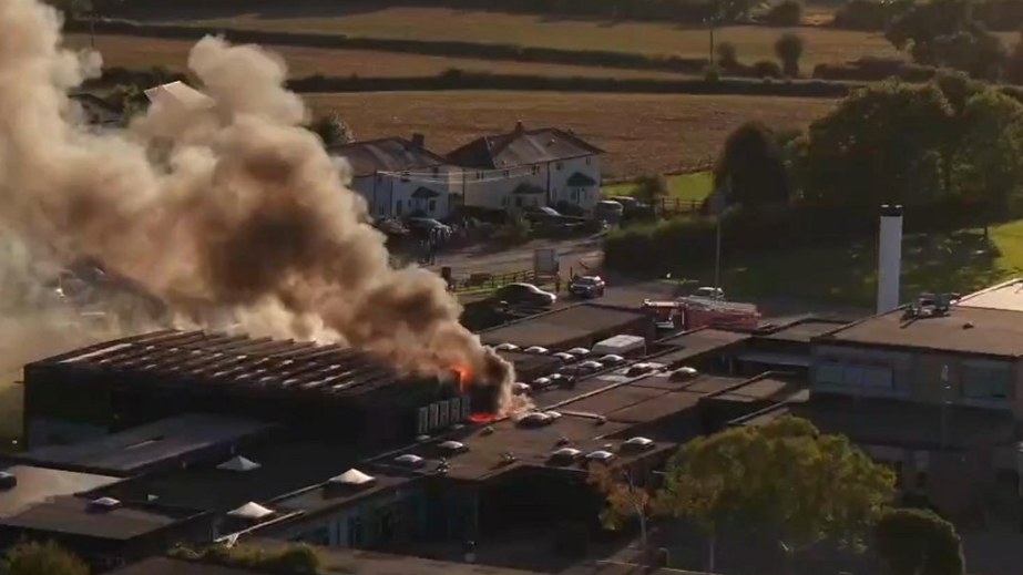 A drone picture shows orange flames and a large, thick plume of black and grey smoke is rising from the roof of a school building. There are houses and card parked nearby, with green fields and trees in the background