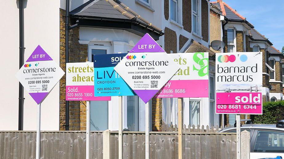 A row of estate agent 'for sale' and 'sold' signs in front of a fence on a residential street in London.