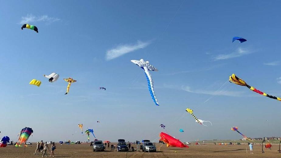 Colourful kits fly in the air above cars and people on a beach. 