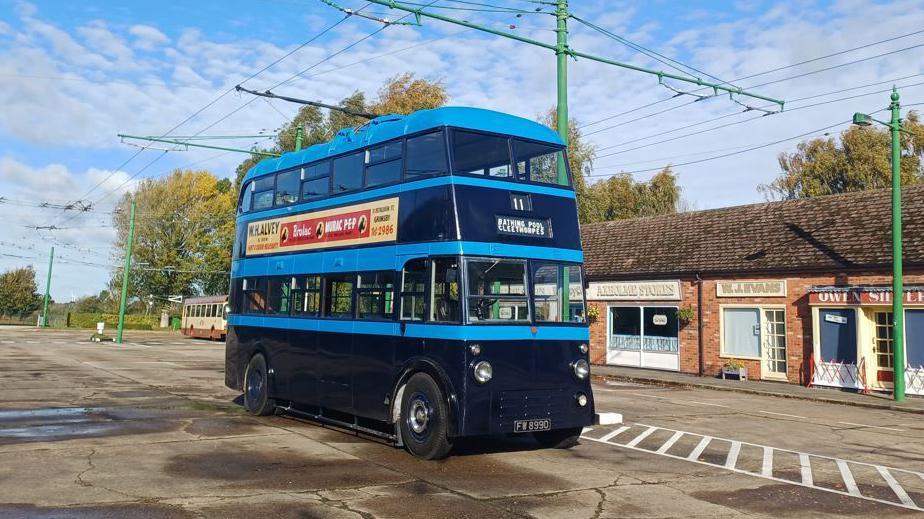 A restored blue and black double-decker vintage trolleybus parked in the middle of a car park next to small buildings. It has "11" and "Cleethorpes" on the front.