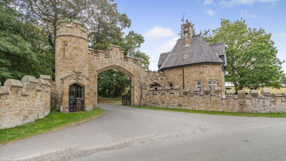 A castellated stone arch entrance to the property with a small stone building to the right. A sign says Gyrn Castle.