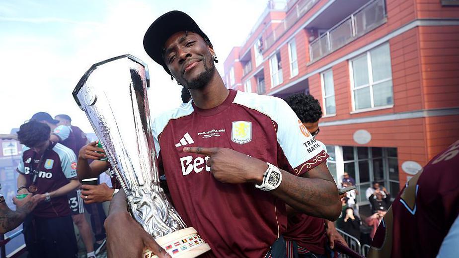 Tammy Abraham holds the Europa League trophy on top of a bus in Aston Villa's parade while he smiles and points at it