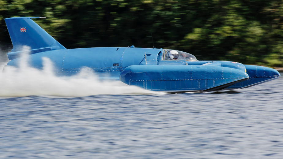 Bluebird, a blue metal hydroplane, crosses Loch Fad with spray shooting up in its wake.
