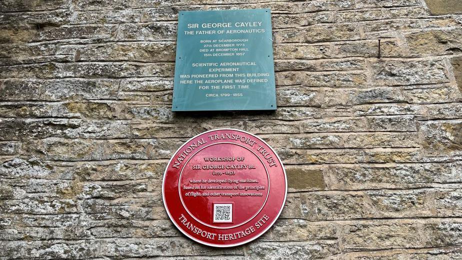 A green plaque and a red plaque on a brick wall.
The green plaque says "Sir George Cayley, the Father of Aeronautics".
The red plaque is from the Transport Heritage Trust and says "Workshop of George Cayley".