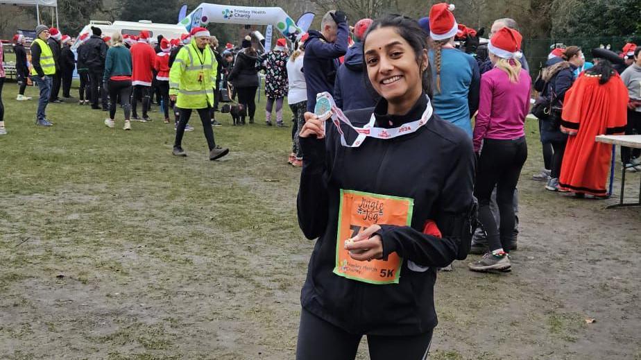 Vasuki Sellathurai is wearing a black top and running leggings. Her long dark hair is tied back and she is smiling at the camera. She has an orange running number pinned to her front. She is holding up a medal. Behind her you can see other runners. Some are wearing santa hats. She is standing on muddy grass. 