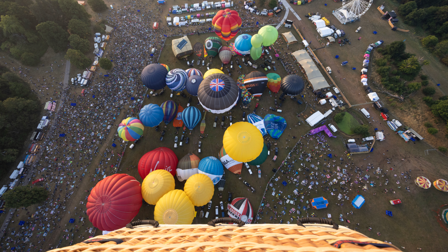 The tops of dozens of colourful hot air balloons in various states of inflation seen from high above. The wicker basket of the balloon the photographer is inside is visible in the bottom of the image. There are large crowds on the ground.