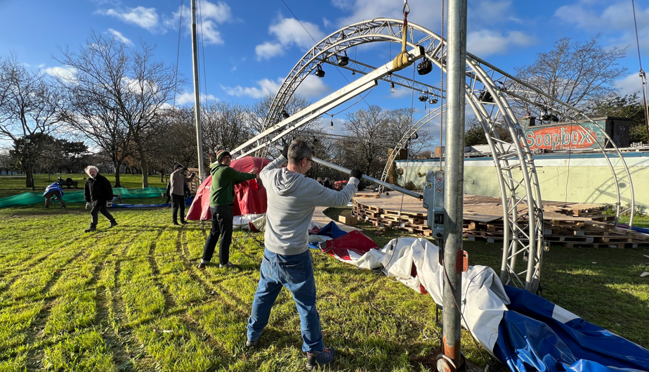 A large tent's metalwork is brought down by four workers in a park with a canvas lying on the ground and a building in the background with the words Soapbox.