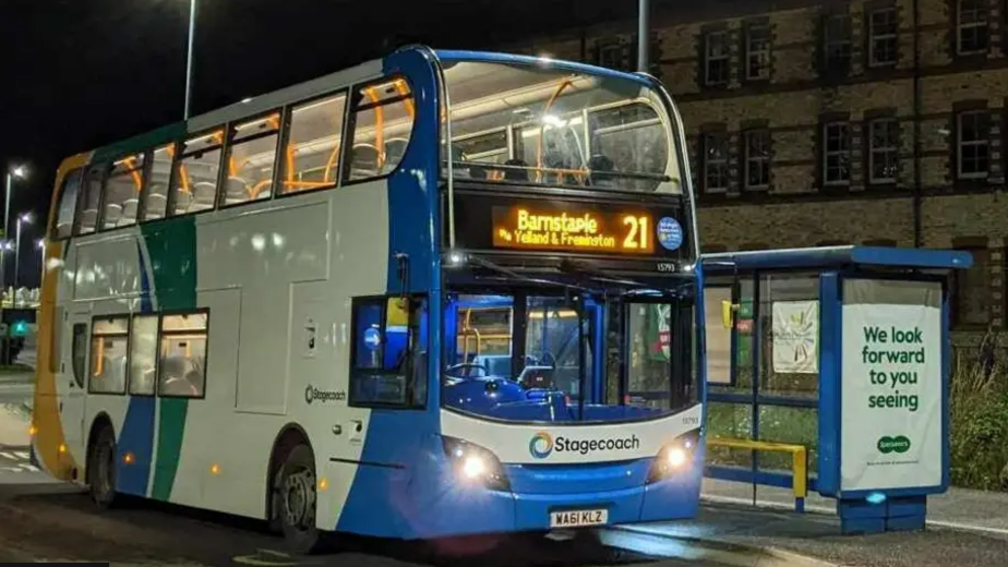 A double-decker bus, pictured at night at a deserted bus stop. Operated by Stagecoach, the bus is painted blue, white, green and yellow and has  "Barnstaple" on its destination board.