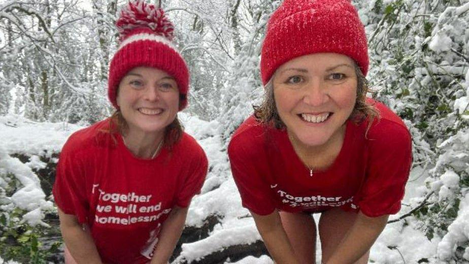 Two women in red bobble hats and t-shirts pose in the snow 