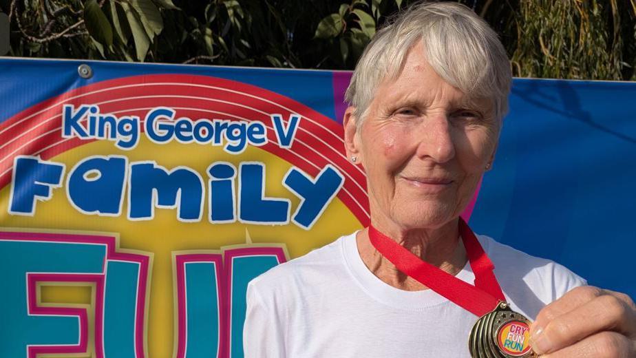 A woman with short grey hair is wearing a white top and holding up a medal which says Cry Fun Run on it. Behind her is a colourful banner which says King George V Family Fun.