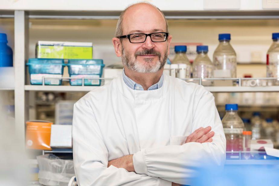 Man with grey beard and black-rimmed glasses, wearing a white medical jacket, arms folded, smiling, in a laboratory setting.