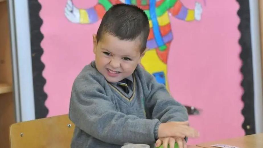 Kyran has short brown hair and is wearing a grey v-neck school jumper with a black and yellow stripe around the collar. He is wearing a grey shirt beneath it and a black tie with a white and yellow stripe. He is sitting at a wooden desk on a wooden chair. Behind him is a pink noticeboard with a colourful display and a black border. You can also see a wooden shelving unit behind him.