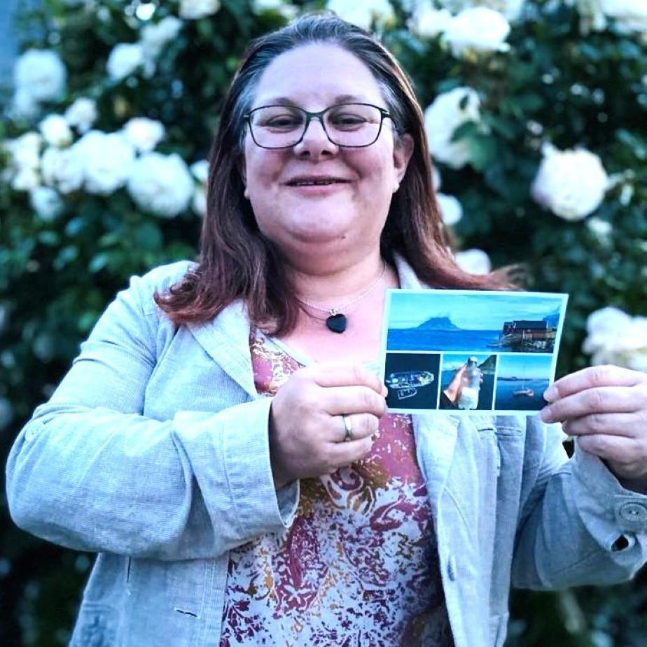 Woman - Alaina Beresford - smiling at camera and holding a postcard, which shows a message in a bottle she sent 31 years ago, after it was found.