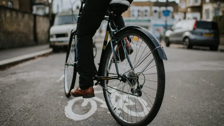 A bicycle is on a road going over a white bicycle sign on the floor. The person riding the bike is wearing black trousers and brown shoes. Cars are parked on either sides of the road. 
