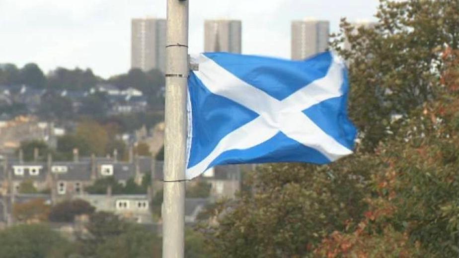 A blue and white saltire flag fluttering on a lamppost, with the city of Aberdeen in the background.
