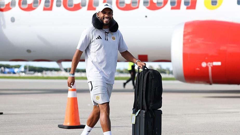 Keni Gorre in a white Curaco polo shirt and shorts,m baseball cap and wearing a neck pillow, walks wheeling black suitcases in front of an aeroplane.