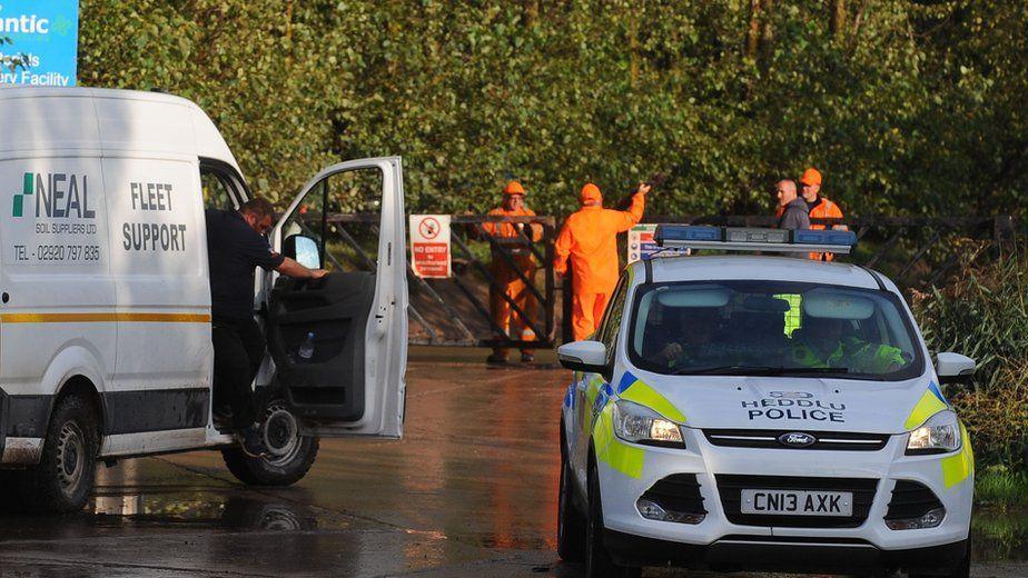 Police and a Neal soils van at the scene of an accident wit worker in orange jump suit manning gates