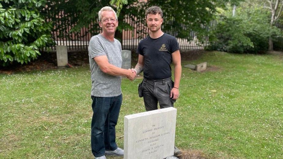 A man with short grey hair wearing a grey patterned t-shirt and blue jeans stood next to a gravestone in a graveyard. He is shaking hands with a man with short brown hair who is wearing a black t-shirt and black trousers. The grass is green and green trees can be seen in the background.