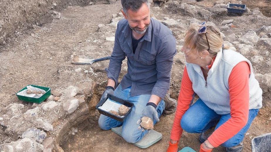 Two archaeologists on the ground in a dig with the foundations of old walls around them. The walls look like pieces of old plaster with bits of colour on.