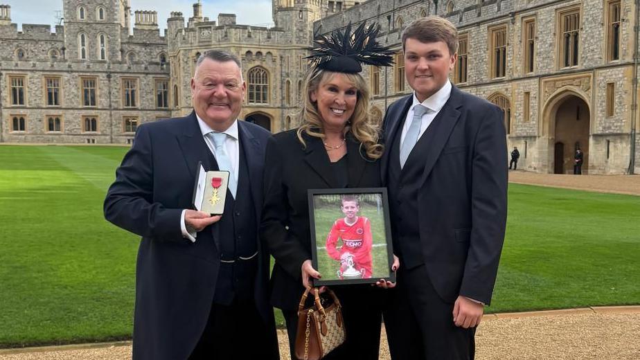 A middle-aged couple and a younger man - all wearing smart dark-coloured suits - pose for a photograph at Windsor Castle. The older man is holding an OBE and the woman is holding a framed photo of a boy dressed in a red football kit. 