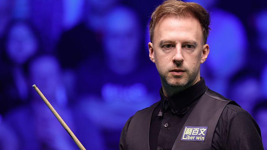 Judd Trump in black shirt and waistcoat carrying a snooker cue