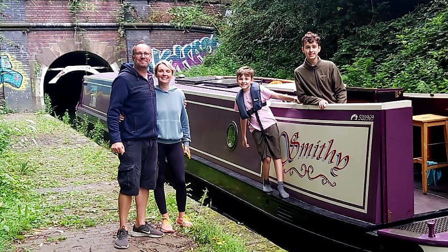 A man and a woman stand on a canal path smiling, as two boys stand on a purple canal boat called Smithy, in front of a canal bridge with colourful grafitti on it