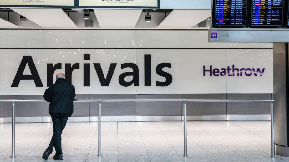 Man waiting in front of a large 'Arrivals' sign at Heathrow airport