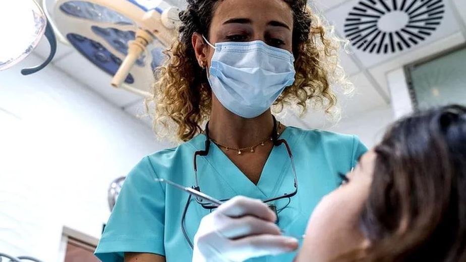 Images shows a black, young female dentist with blonde and pale brown curly hair checking a woman with dark hair's teeth