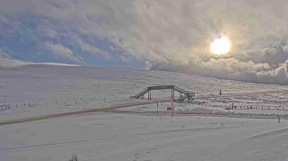 Snaefell Mountain covered in snow, with some clearance of the carriageway. There is a footbridge over the road and the sun can partially be seen behind some clouds.