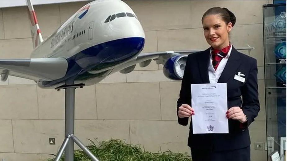 A woman in an air steward's uniform stands next to a statue of a British Airways aeroplane. The woman has dark brown hair and she is wearing a navy blue skirt suit, a white shirt, and a red and white scarf. She is also holding a certificate. The aeroplane statue is mostly white, with red and blue designs on it.