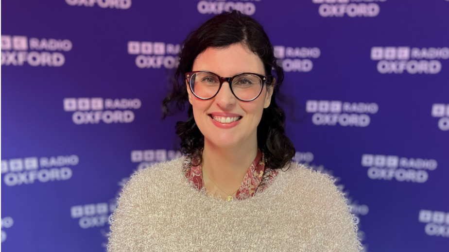 A photo of Layla Moran MP in front of purple branding for Radio Oxford. She is wearing dark rimmed glasses and a fluffy beige jumper.