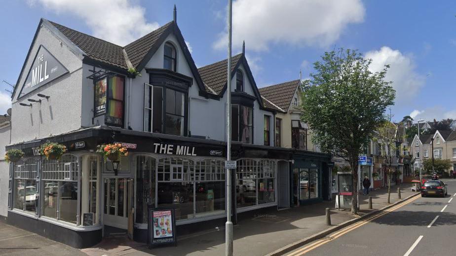 An exterior shot of the Mill, a white building with a big windows wrapping around the ground floor. There are bay windows on the first floor. There is a stretch of pavement outside the pub with a tree and bollards.
