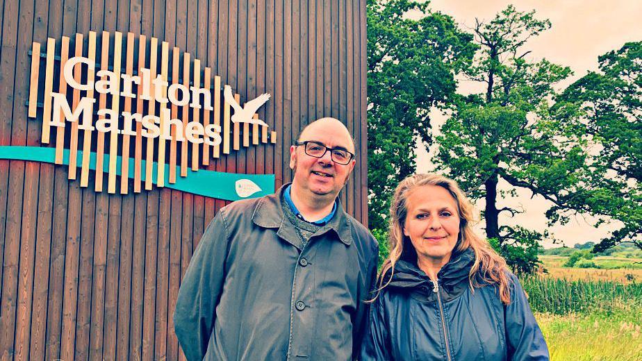 Dean Parkin and Naomi Jaffa smile at the camera as they are photographed outside. They are dressed in dark raincoats and are standing in front of building with a sign that reads: "Carlton Marshes." 