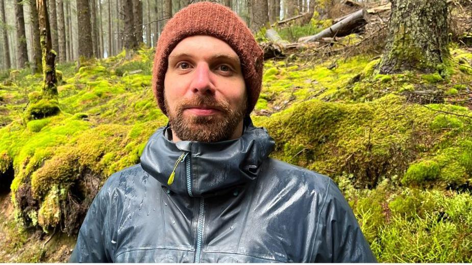 Ffred Kehoe mewn coedwig ble mae mae mwsogl yn gorchuddio'r tir