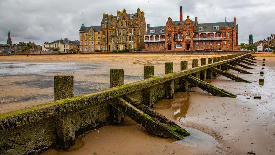 View of Portobello Beach in Edinburgh, Scotland, featuring a wooden groyne leading towards historic Victorian-style buildings under a cloudy sky