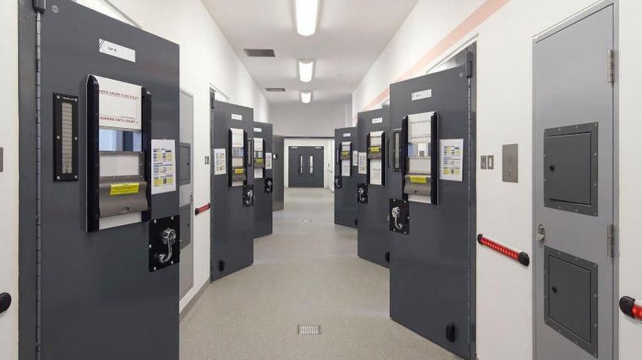 A corridor with police cells either side. The dark grey cell doors have been opened and double doors are visible at the end of the corridor.