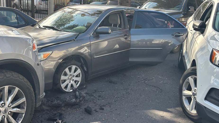A Toyota Camry is parked between several other cars with its rear driver side door open and tires blown out.