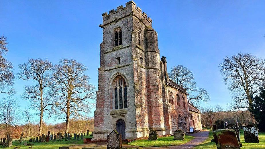 A church with a square tower sits surrounded by gravestones and some bare trees against a blue sky.