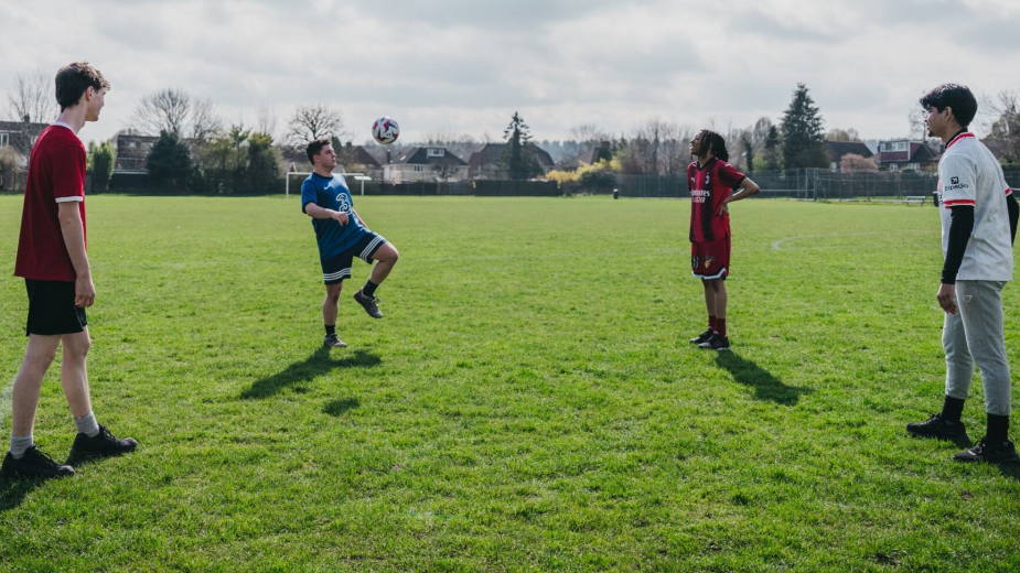 Four boys standing on a football pitch in sports gear, kicking a football around together. There are houses and trees in the background.